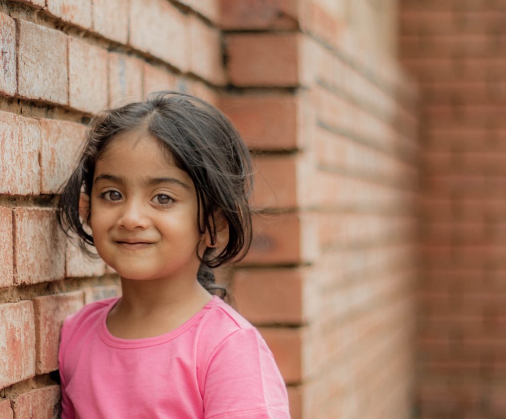 little girl standing by a brick wall with a small smirk. 