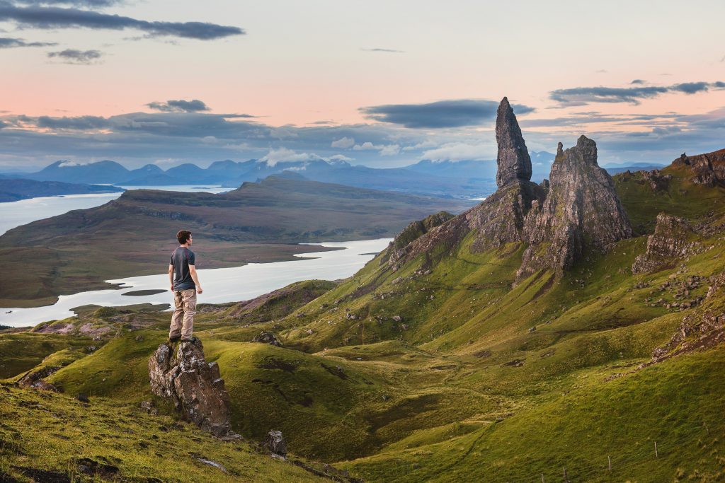 The Highlands in Scotland shows a man standing on a rock with green fields and mountain sticking out; lake in the background