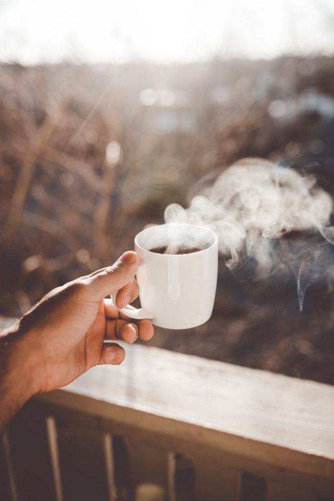 A man's hand with a cup of hot coffee, steaming coming out of it, out on a balcony.