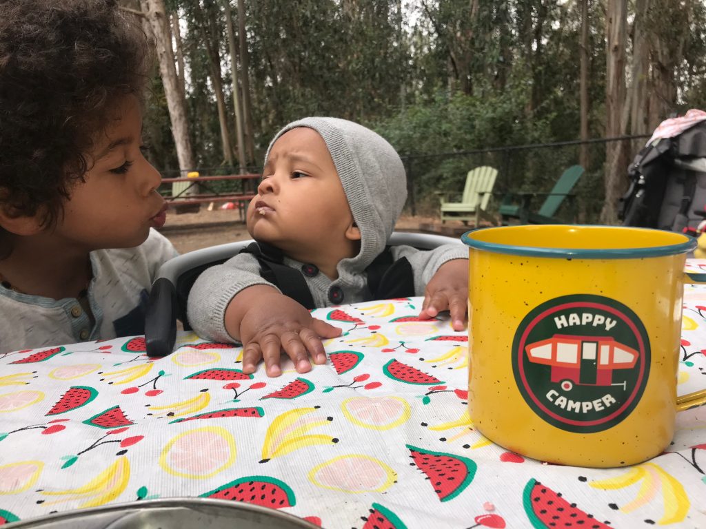 Baby and his big brother looking at each other at a picnic table while camping. 