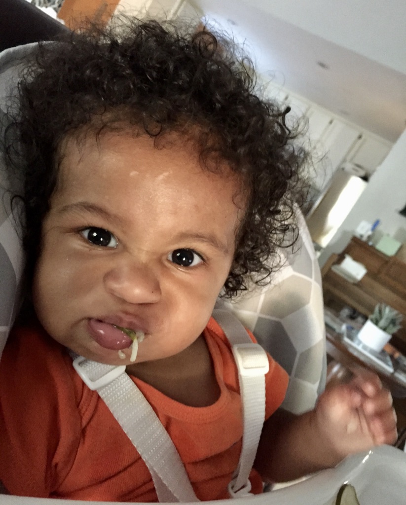 A toddler in a high chair pushing food out with his tongue. 