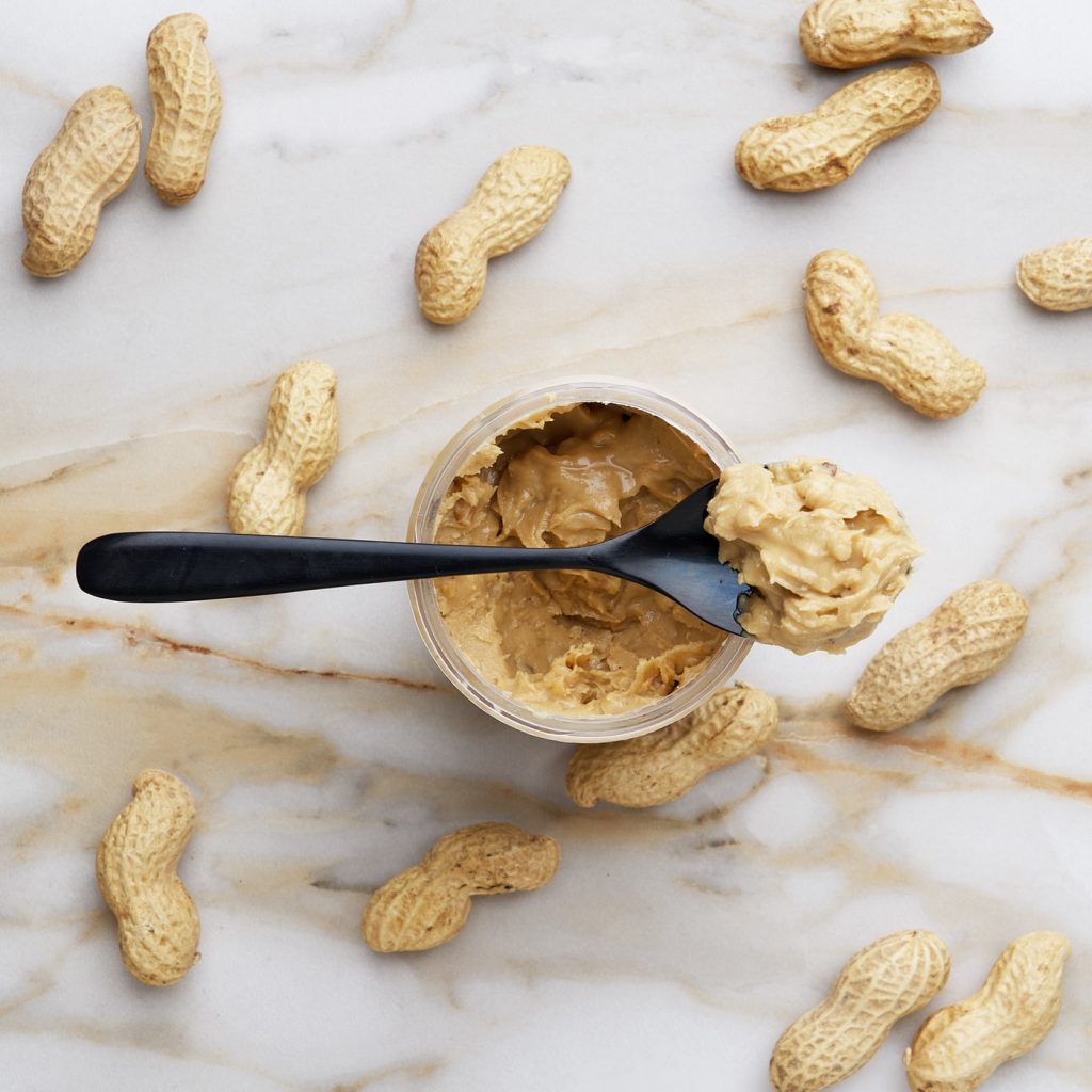 peanut butter in a jar with a spoon sitting on a countertop with hold peanuts around it.